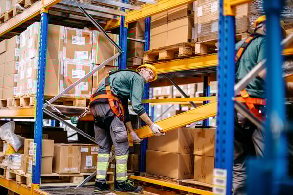 Innovative Workers' Compensation: A warehouse male worker wearing safety gear is lifting a beam while secured to a harness, surrounded by shelves stacked with boxes.