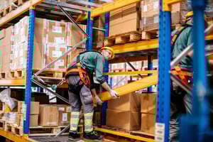 Innovative Workers' Compensation: A warehouse male worker wearing safety gear is lifting a beam while secured to a harness, surrounded by shelves stacked with boxes.
